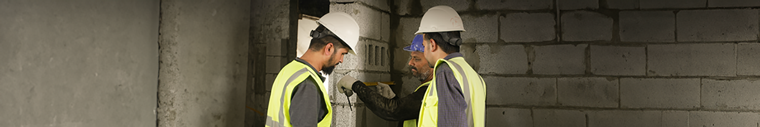 3 men ins hi-vis vests inspecting a wall of a building with a measuring tape